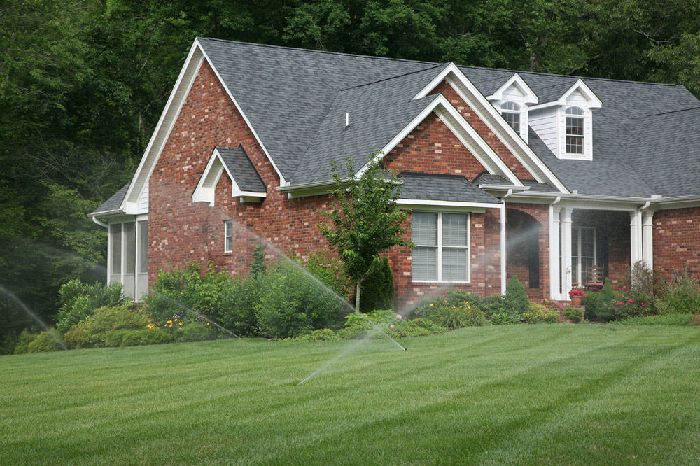 Sprinklers Watering Green Lawn With Brick House In Background